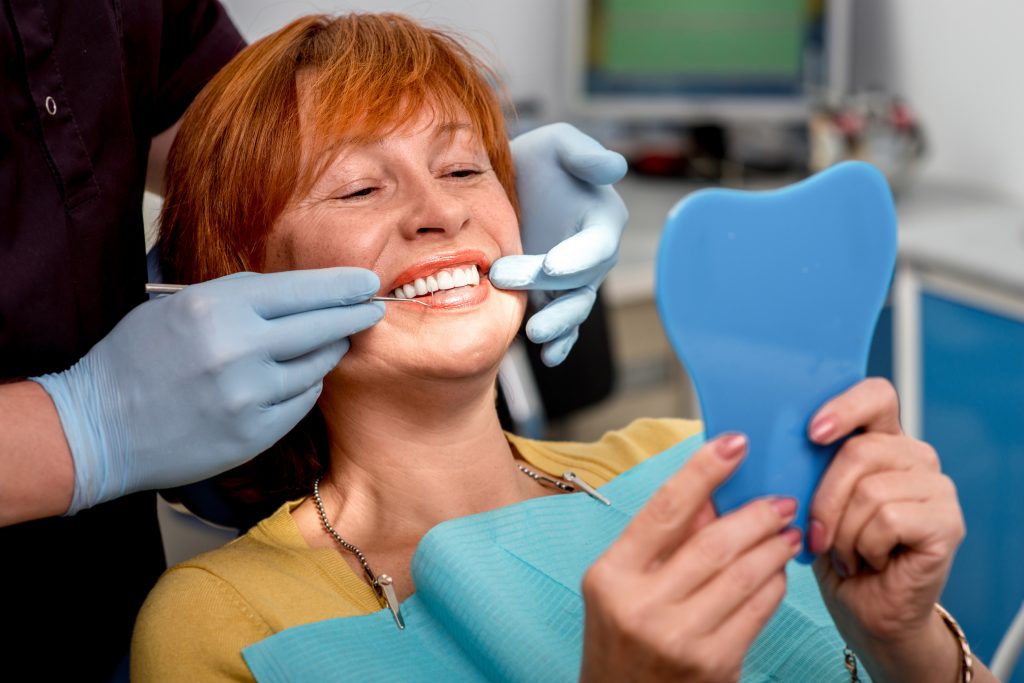 Smiling senior woman with new dentures sitting in the dental office and looking at the mirror